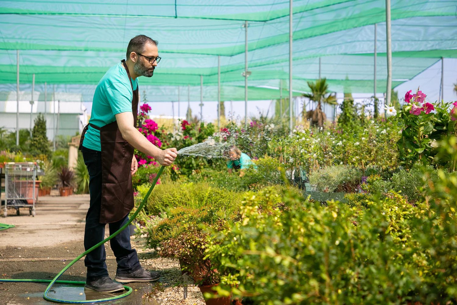 bearded-man-holding-hose-standing-watering-plants-unrecognizable-blurred-colleague-growing-flowers-two-gardeners-wearing-uniform-working-hothouse-gardening-activity-summer-concept_1